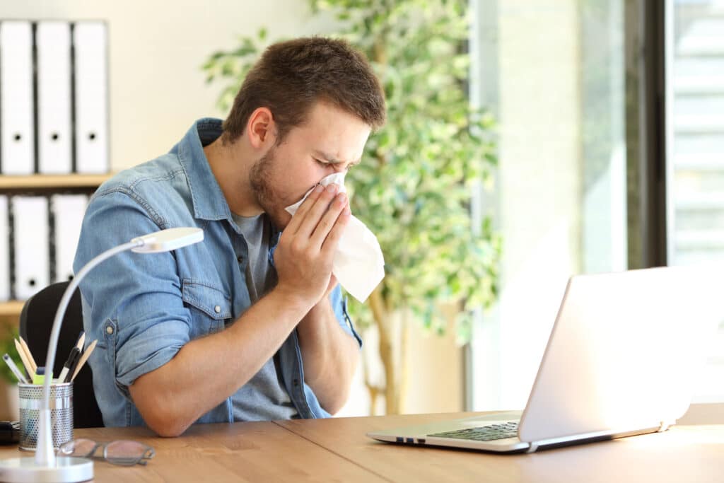 Man blowing nose into a tissue while working in his home office